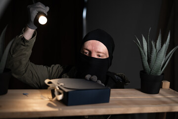 thief in black balaclava stealing dark box with money from bookshelf.
