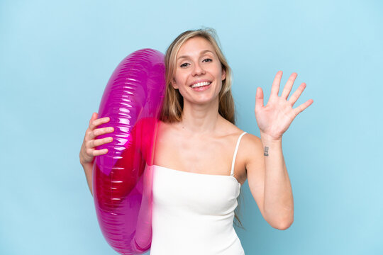 Young blonde woman in swimsuit holding inflatable donut isolated on blue background saluting with hand with happy expression - Powered by Adobe