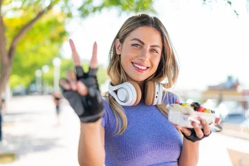 Fotobehang Young pretty sport woman holding a bowl of fruit at outdoors smiling and showing victory sign © luismolinero