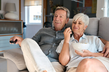 Happy senior couple watching tv and eating popcorn on sofa