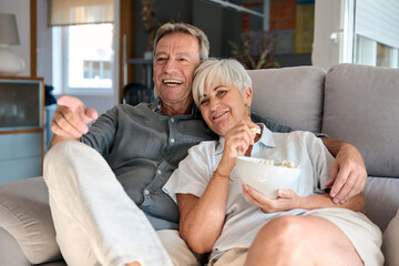 Obraz premium Happy senior couple watching tv and eating popcorn on sofa