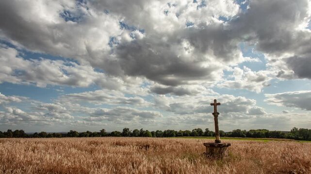 Solitary stone cross in golden wheat field beneath dramatic sky with heavy clouds. Peaceful and spiritual atmosphere of countryside near Torgny in Southern Belgium.