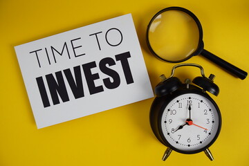 Time to Invest text with magnifying glass and alarm clock flat lay on yellow background
