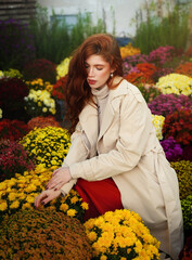 Beautiful red-haired young woman in beige trench coat at city flower market among colorful autumn chrysanthemums. Autumn season.