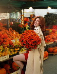 Beautiful red-haired young woman with orange bouquet of autumn flowers in her hands walks through the city vegetable market. Autumn and Halloween season.