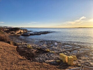 The craggy and jagged rocky coastline attracts incoming waves as setting sunlight reflections make the rippled water glow. The silhouette of a wharf points towards cranes on the horizon.