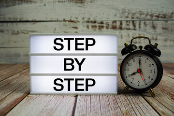 Step by Step text on light box with alarm clock on wooden background