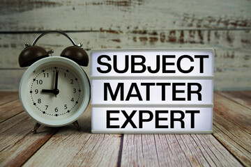 Subject Matter Expert text on light box with alarm clock on wooden background