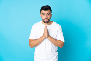 Caucasian man over isolated blue background keeps palm together. Person asks for something