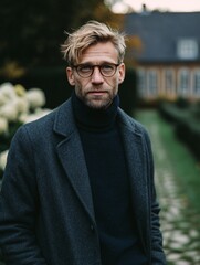Obraz premium Bearded man in glasses and coat stands confidently outside, capturing a fashionable autumn vibe in a serene setting.