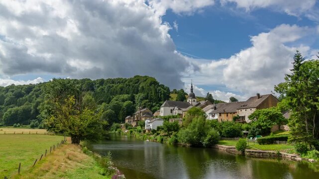 Picturesque village nestled by calm river, surrounded by lush greenery and rolling hills. Time lapse with vibrant sky and fluffy clouds.