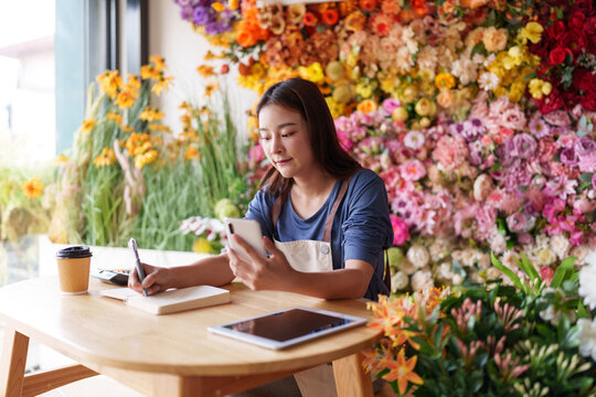 Asian woman small business owner manages artificial flower shop sales marketing using smartphone writing orders creative floral studio vibrant retail store