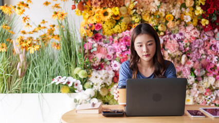 Asian woman small business owner working laptop computer managing artificial flower shop orders customer service vibrant floral decor retail entrepreneurship