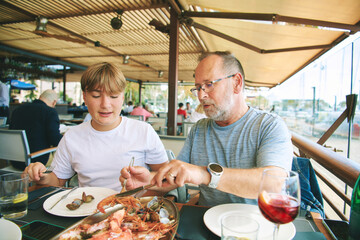 Outdoor portrait of father and teenager son eating seafood in seaside restaurant