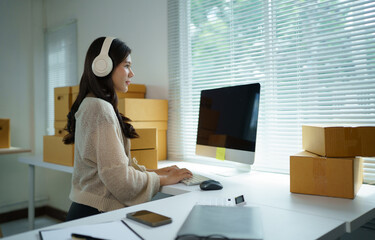 Young Asian Woman Entrepreneur Wearing Headphones Working on Computer in Home Office with Cardboard...