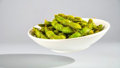 A white ceramic bowl of seasoned green edamame pods levitating over a grey surface.