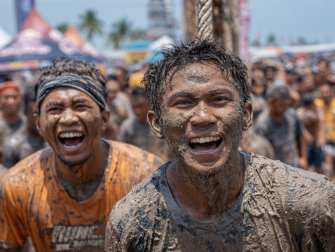 Participants grinning and shouting in delight during a slippery pole-climbing contest at the Perlis Water Festival, bodies covered in mud, playful struggle to reach the top