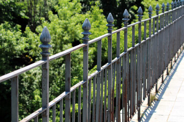 A linear decorative wrought iron fence features vertical bars and spear-shaped finial tops, casting...