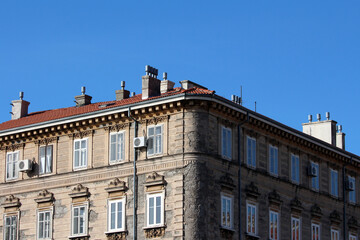 Obraz premium A multi-story European-style stone building with ornate window frames, red-tiled roof, numerous rooftop chimneys, and decorative exterior details under a clear blue sky