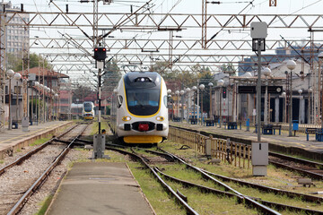 A sleek, modern electric train with a yellow and white livery approaches an urban railway platform, surrounded by signal lights and complex overhead wiring at a city train station