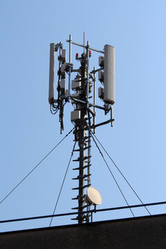 High-rise cell tower featuring vertical panel antennas, a parabolic dish, support cables, and exposed wiring, standing against a clear blue sky for urban telecommunications infrastructure