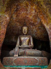 A Buddha statue in a meditation pose, seated inside a natural cave. The sculpture shows the...