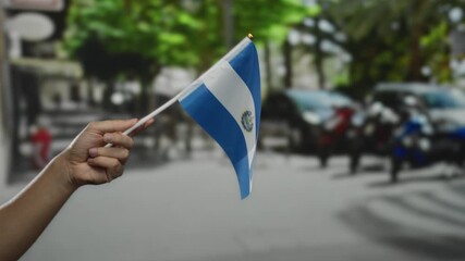 Hand holds el salvador flag outdoors on a sunny day in a lively city street, representing national pride, culture, and identity.