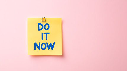 A yellow sticky note with blue lettering on it reads 'DO IT NOW' against a soft pink backdrop; a reminder of urgency, productivity, and immediate action.