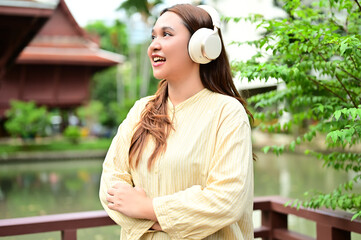 Asian Woman Smiling and Enjoying Music with Wireless Headphones Outdoors by Pond.