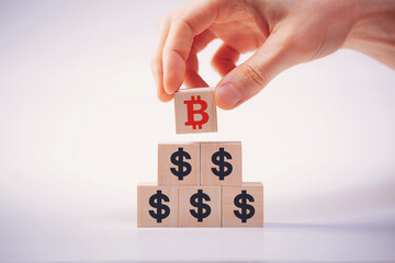 A hand places a wooden cube with a Bitcoin sign on top of a dollar pyramid, symbolizing the rise of cryptocurrency. Symbolizes the transition of the world currency to digital money, the future of bloc