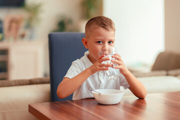 Cute 5-year-old boy having breakfast at home, eating porridge with water. Natural light, healthy food