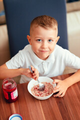 Cute 5-year-old boy having breakfast at home, eating porridge with jam.