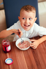 Cute 5-year-old boy having breakfast at home, eating porridge with jam.