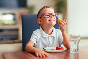 Cute boy in glasses eating vegetables at home