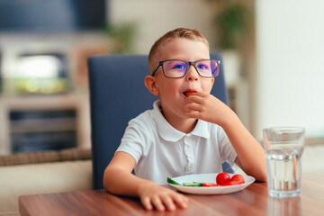 Cute boy in glasses eating vegetables at home
