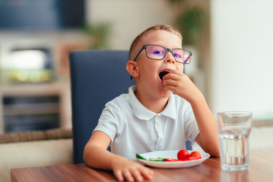 Cute boy in glasses eating vegetables at home
