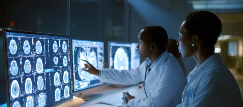 A male and female doctor are examining several brain scans on a computer screen in a dark office. The male doctor points to a particular scan on the monitor, while the female doctor listens intently