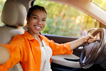 Black young woman smiling for selfie in the car, one hand on wheel. Confident and modern portrayal...