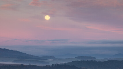 Full moon glows above a misty landscape, casting a soft, ethereal light over the scene.
