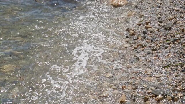 Sunlit mediterranean beach scene captures gentle waves meeting a pebbled shoreline outdoors, emphasizing natural beauty and relaxation under a clear sky.