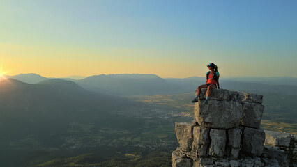 Fototapeta premium Hiker triumphantly sits on a rocky peak at sunrise, overlooking a vast valley. Environmental portrait captures achievement, endurance, and freedom, with golden light symbolizing perseverance.