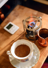 A traditional copper Turkish coffee pot filled with beans rests on a digital scale alongside a kettle on a wooden counter, evoking a scene of artisanal coffee preparation.