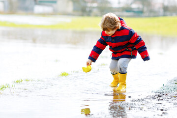 Happy little kid boy in yellow rain boots playing with paper ship boat by huge puddle on spring or...