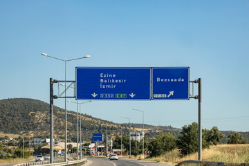 Highway signs showing directions to Ezine, Balıkesir, İzmir and Bozcaada, with countryside view under clear blue sky, Turkey