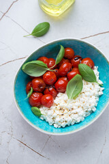 Cottage cheese caprese with roasted tomatoes and basil served in a turquoise bowl, vertical shot on a white stone surface, elevated view