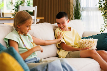 Brother and sister sharing popcorn while watching tv on sofa at home