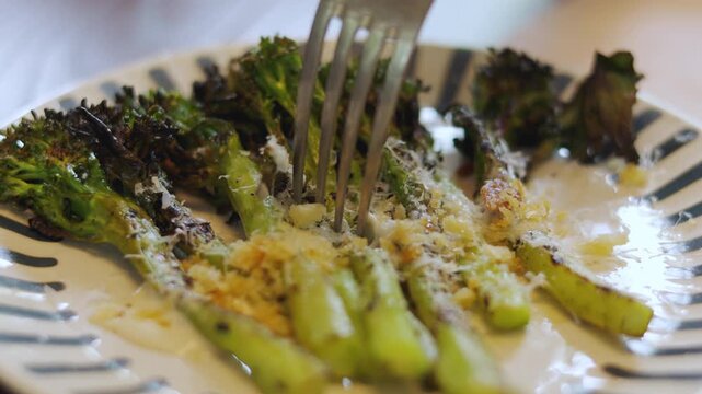 Close-up of roasted broccolini with parmesan and crunchy crumbs on ceramic plate. Selective focus. Fork pressing vegetables. Healthy, vegetarian, flavorful dish captured in natural warm lighting.