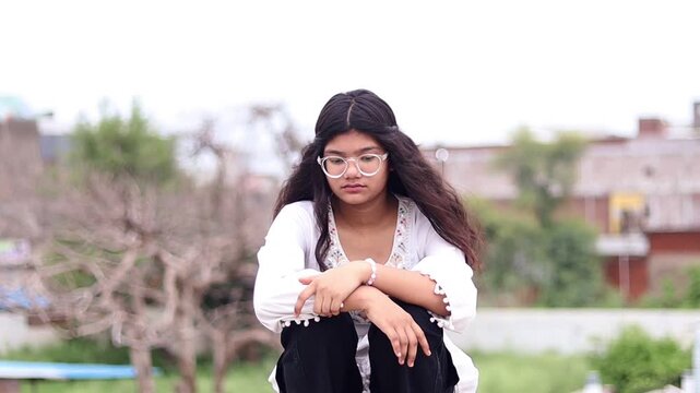 Cinematic side-panning clip of a 15-year-old girl wearing spectacles, sitting alone with a sad expression, lost in thought, in a natural outdoor setting with soft light