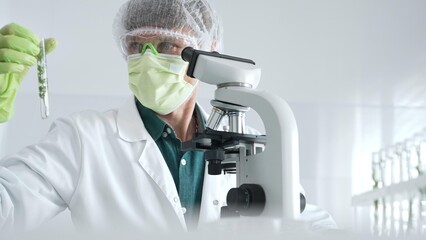 Adult male scientist wearing green protective gear looking at plant samples in test tubes and using...