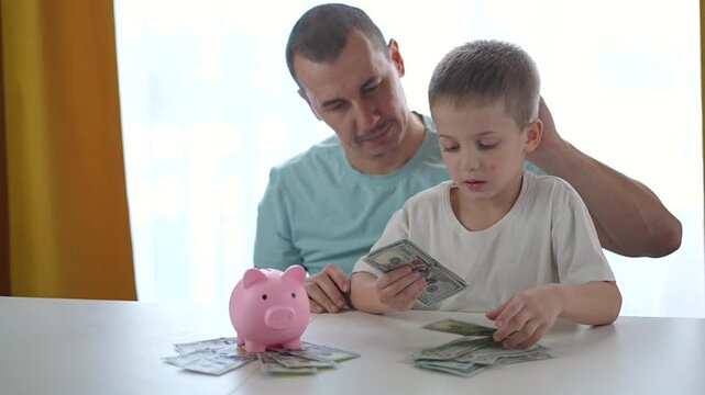 Boy counts money while father explains saving again and again. Father helps boy handle dollar bills. Saving taught by father with clear finance steps. Boy repeats saving with father support.
